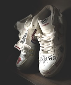 A pair of white sneakers with various logos and text designs, including team names and brand labels. They have a high-top style and intricate detailing on their surface, showcasing a blend of sporty and trendy aesthetics.