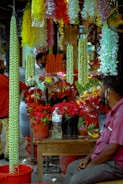 A bustling market scene with vibrant floral displays. Several bright garlands of flowers hang from above, predominantly featuring green, yellow, and white colors. Buckets filled with red roses are prominently placed on a wooden table. In the foreground, a man in a purple shirt is seated, looking towards the flower arrangements. Other figures in the background appear to be engaging in conversations or customer interactions.