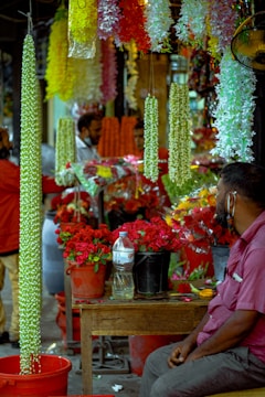 A bustling market scene with vibrant floral displays. Several bright garlands of flowers hang from above, predominantly featuring green, yellow, and white colors. Buckets filled with red roses are prominently placed on a wooden table. In the foreground, a man in a purple shirt is seated, looking towards the flower arrangements. Other figures in the background appear to be engaging in conversations or customer interactions.