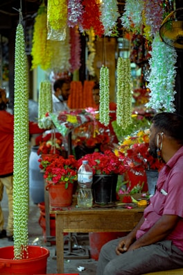 A bustling market scene with vibrant floral displays. Several bright garlands of flowers hang from above, predominantly featuring green, yellow, and white colors. Buckets filled with red roses are prominently placed on a wooden table. In the foreground, a man in a purple shirt is seated, looking towards the flower arrangements. Other figures in the background appear to be engaging in conversations or customer interactions.