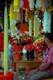 A bustling market scene with vibrant floral displays. Several bright garlands of flowers hang from above, predominantly featuring green, yellow, and white colors. Buckets filled with red roses are prominently placed on a wooden table. In the foreground, a man in a purple shirt is seated, looking towards the flower arrangements. Other figures in the background appear to be engaging in conversations or customer interactions.