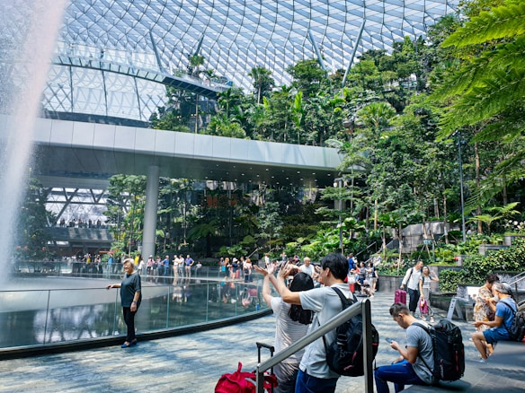 A large indoor rainforest with a glass dome ceiling, featuring a waterfall surrounded by lush greenery. A group of people are taking photos and walking around the area. The environment is modern and bustling with visitors.