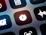 A close-up view of a mobile device screen displaying application icons in a grid layout. Visible icons include a stopwatch, a sound wave, an alarm clock, and other utility symbols. The design is sleek with icons having a minimalist, monochromatic style against a dark background.