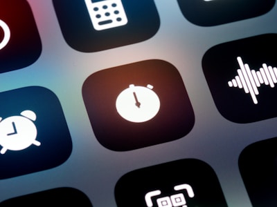 A close-up view of a mobile device screen displaying application icons in a grid layout. Visible icons include a stopwatch, a sound wave, an alarm clock, and other utility symbols. The design is sleek with icons having a minimalist, monochromatic style against a dark background.