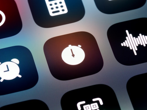 A close-up view of a mobile device screen displaying application icons in a grid layout. Visible icons include a stopwatch, a sound wave, an alarm clock, and other utility symbols. The design is sleek with icons having a minimalist, monochromatic style against a dark background.