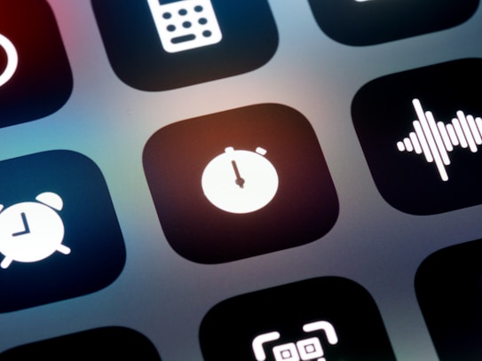 A close-up view of a mobile device screen displaying application icons in a grid layout. Visible icons include a stopwatch, a sound wave, an alarm clock, and other utility symbols. The design is sleek with icons having a minimalist, monochromatic style against a dark background.