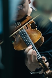 A person is intently playing a violin, with a focus on the hand and strings. The violinist appears to be in deep concentration, and the scene is partially obscured by a blurred object in the foreground.