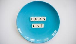 A blue plate with the words 'BURN FAT' spelled out in letter tiles placed in the center. The background is white which contrasts with the blue plate.