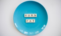 A blue plate with the words 'BURN FAT' spelled out in letter tiles placed in the center. The background is white which contrasts with the blue plate.