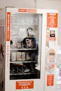 A vending machine with a glass front displaying various packaged snack items. The machine has labels in multiple languages including Japanese and Indonesian. It features a keypad for input and a currency slot for transactions, with instructions available on bright orange panels.