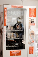 A vending machine with a glass front displaying various packaged snack items. The machine has labels in multiple languages including Japanese and Indonesian. It features a keypad for input and a currency slot for transactions, with instructions available on bright orange panels.