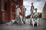 Metal sculptures of abstract human figures arranged in front of a brick building. The figures appear elongated and stylized, each with unique geometric designs and forms. The sculptures are set on a cobblestone path, with overcast skies and nearby trees in the background.