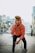A young man wearing a pink hooded sweatshirt with a 'Supreme' logo stands in an urban street setting. He has styled hair and black pants, striking a dynamic pose with one hand near his face. The background features a blurred cityscape with buildings and a few parked cars.
