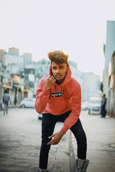 A young man wearing a pink hooded sweatshirt with a 'Supreme' logo stands in an urban street setting. He has styled hair and black pants, striking a dynamic pose with one hand near his face. The background features a blurred cityscape with buildings and a few parked cars.