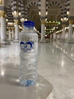 A clear plastic water bottle with a blue label and cap is set on a glossy marble surface. In the background, the interior of a grand building features tall, ornate columns with intricate patterns and soft lighting from gold and white fixtures.