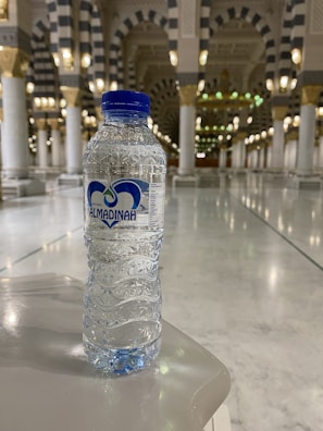 A clear plastic water bottle with a blue label and cap is set on a glossy marble surface. In the background, the interior of a grand building features tall, ornate columns with intricate patterns and soft lighting from gold and white fixtures.