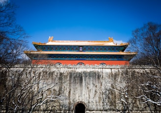 A traditional Chinese building with ornate architectural details stands atop a large stone wall. The structure features vibrant blue and red hues with a golden roof, surrounded by bare trees under a clear blue sky.