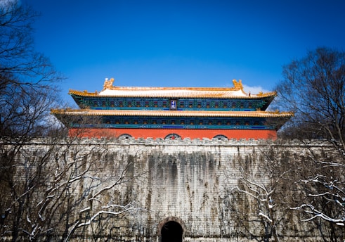 A traditional Chinese building with ornate architectural details stands atop a large stone wall. The structure features vibrant blue and red hues with a golden roof, surrounded by bare trees under a clear blue sky.