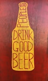 A graphic stylized as a beer bottle with the text 'Good People Drink Good Beer' written in bold, uppercase letters. The design features a mustard yellow bottle shape against a textured, dark reddish-brown background.