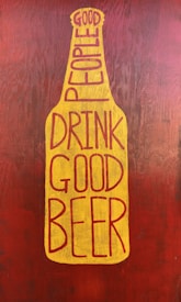 A graphic stylized as a beer bottle with the text 'Good People Drink Good Beer' written in bold, uppercase letters. The design features a mustard yellow bottle shape against a textured, dark reddish-brown background.