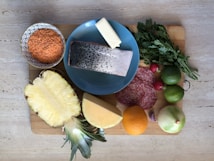 A variety of fresh ingredients are placed on a wooden cutting board. There is a bowl of red lentils, a slice of fish with skin on a blue plate, a block of butter, fresh parsley, sliced salami, a halved pineapple, a cut of cheese, an orange, and several vegetables including radishes, limes, a garlic clove, and an onion.