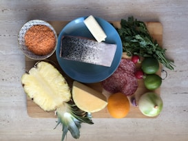 A variety of fresh ingredients are placed on a wooden cutting board. There is a bowl of red lentils, a slice of fish with skin on a blue plate, a block of butter, fresh parsley, sliced salami, a halved pineapple, a cut of cheese, an orange, and several vegetables including radishes, limes, a garlic clove, and an onion.