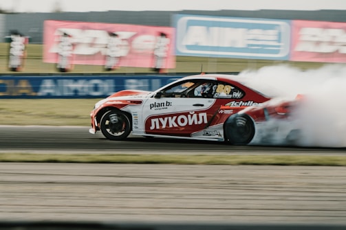 A sports car drifts at high speed on a racetrack, emitting thick clouds of white smoke from its back tires. The car is predominantly white and red, with various sponsor logos displayed across its body. Background signage and blurred motion suggest a fast-paced, dynamic environment.