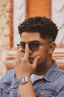 A person wearing glasses and a denim jacket focuses intently, partially covering their face with one hand. They have curly hair and wear a digital watch on their wrist. The background features a textured wall with geometric patterns and earthy tones.