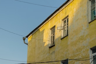 A sunlit yellow building wall with windows, some of which are open, and several electrical wires crossing in front. The paint is slightly weathered, giving the facade a rustic appearance.