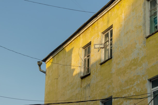 A sunlit yellow building wall with windows, some of which are open, and several electrical wires crossing in front. The paint is slightly weathered, giving the facade a rustic appearance.