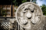 A detailed stone carving featuring intricate designs with mythical creatures and floral patterns. The carving is set against a blurred background that includes a traditional building with wooden and brick elements, partially obscured by greenery.