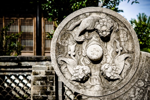 A detailed stone carving featuring intricate designs with mythical creatures and floral patterns. The carving is set against a blurred background that includes a traditional building with wooden and brick elements, partially obscured by greenery.