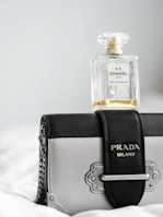 A bottle of Chanel No. 5 perfume rests atop a stylish Prada Milano handbag. The perfume bottle is clear with a classic design, while the handbag features a combination of black and metallic elements with a chain strap.