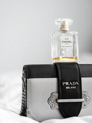 A bottle of Chanel No. 5 perfume rests atop a stylish Prada Milano handbag. The perfume bottle is clear with a classic design, while the handbag features a combination of black and metallic elements with a chain strap.