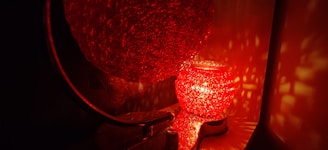 A glowing red textured lamp casting intricate light patterns on the surrounding surfaces. The lamp, which appears to be spherical, emits a warm and inviting light, creating a cozy ambiance. The light reflections add a mesmerizing effect to the overall scene.
