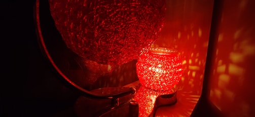 A glowing red textured lamp casting intricate light patterns on the surrounding surfaces. The lamp, which appears to be spherical, emits a warm and inviting light, creating a cozy ambiance. The light reflections add a mesmerizing effect to the overall scene.