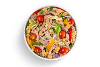A bowl filled with a colorful mixture of cooked pasta, chunks of tuna, cherry tomatoes, sliced red onions, yellow and green bell peppers, corn, and chickpeas. The ingredients are vibrant and fresh, creating a visually appealing dish.