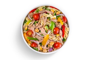 A bowl filled with a colorful mixture of cooked pasta, chunks of tuna, cherry tomatoes, sliced red onions, yellow and green bell peppers, corn, and chickpeas. The ingredients are vibrant and fresh, creating a visually appealing dish.