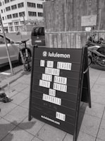 A black sandwich board on a sidewalk in front of a wooden wall and a parked motorcycle. The board has a motivational message from Lululemon encouraging kindness and support for others. In the background, urban buildings and parked cars are visible.