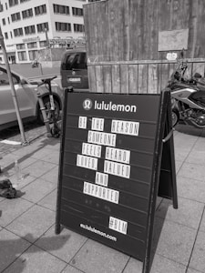 A black sandwich board on a sidewalk in front of a wooden wall and a parked motorcycle. The board has a motivational message from Lululemon encouraging kindness and support for others. In the background, urban buildings and parked cars are visible.