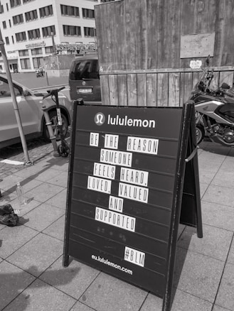 A black sandwich board on a sidewalk in front of a wooden wall and a parked motorcycle. The board has a motivational message from Lululemon encouraging kindness and support for others. In the background, urban buildings and parked cars are visible.
