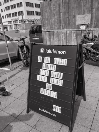 A black sandwich board on a sidewalk in front of a wooden wall and a parked motorcycle. The board has a motivational message from Lululemon encouraging kindness and support for others. In the background, urban buildings and parked cars are visible.