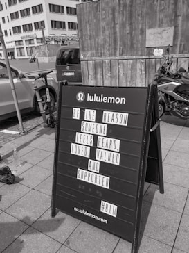 A black sandwich board on a sidewalk in front of a wooden wall and a parked motorcycle. The board has a motivational message from Lululemon encouraging kindness and support for others. In the background, urban buildings and parked cars are visible.