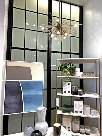 A stylish interior section featuring a modern chandelier with an abstract starburst design hanging from the ceiling. Below, there is a black-framed window providing a geometric backdrop. To the right, a shelving unit displays various decorative items such as ceramics, candles, and plants. On the left, a minimalistic artwork with soft hues of gray and blue hangs above a table showcasing more ceramic vases and a coffee mug.