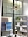 A stylish interior section featuring a modern chandelier with an abstract starburst design hanging from the ceiling. Below, there is a black-framed window providing a geometric backdrop. To the right, a shelving unit displays various decorative items such as ceramics, candles, and plants. On the left, a minimalistic artwork with soft hues of gray and blue hangs above a table showcasing more ceramic vases and a coffee mug.