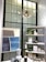 A stylish interior section featuring a modern chandelier with an abstract starburst design hanging from the ceiling. Below, there is a black-framed window providing a geometric backdrop. To the right, a shelving unit displays various decorative items such as ceramics, candles, and plants. On the left, a minimalistic artwork with soft hues of gray and blue hangs above a table showcasing more ceramic vases and a coffee mug.