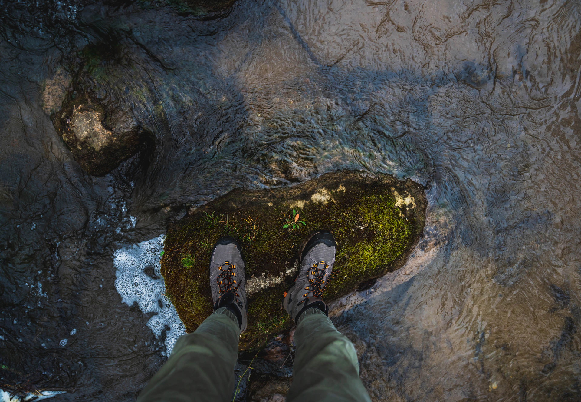 Close-up of sturdy hiking boots stepping over a bubbling mountain stream surrounded by mossy rocks.