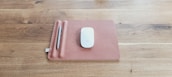 A sleek mouse is placed on a brown rectangular mat which has a slot for holding a silver pen. The mat rests on a wooden surface with a visible grain pattern.