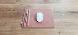 A sleek mouse is placed on a brown rectangular mat which has a slot for holding a silver pen. The mat rests on a wooden surface with a visible grain pattern.