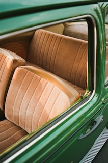 The image captures the interior of a vintage car with tan-colored, cushioned seats featuring vertical stitching. The vehicle's exterior is a rich green color, visible through the open window frame. The overall composition highlights the classic design and texture of the car's seating.